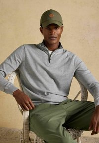 Mann trägt olivgrüne Kappe, hellgrauen Pullover mit Viertel-Reißverschluss und olivgrüne Hose, sitzt entspannt in einem weißen Korbstuhl.