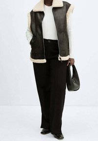 Veste sans manches en peau de mouton noire avec doublure beige clair, fermeture éclair sur le devant et deux poches latérales. Portée par-dessus un haut blanc texturé et associée à un pantalon noir.