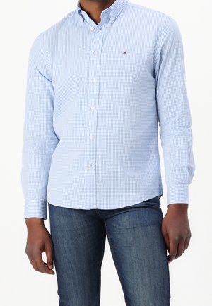 Man wearing light blue checkered button-up shirt with small logo on chest and dark blue jeans against white background.