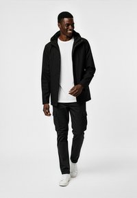 Svart vattentät jacka med huva, vit t-shirt, svarta cargobyxor och vita sneakers. Slät textur, normal passform, avslappnad design.
