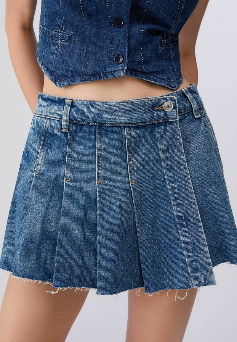 Femme portant une mini-jupe plissée en denim bleu avec une ceinture boutonnée et un gilet en denim court avec des boutons.