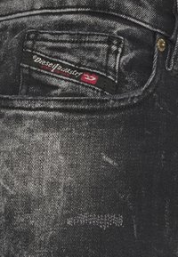 Tejido de mezclilla negra con una textura desgastada. Cuenta con un bolsillo pequeño y una etiqueta roja y negra que dice "Diesel Industry." Detalles en dorado.