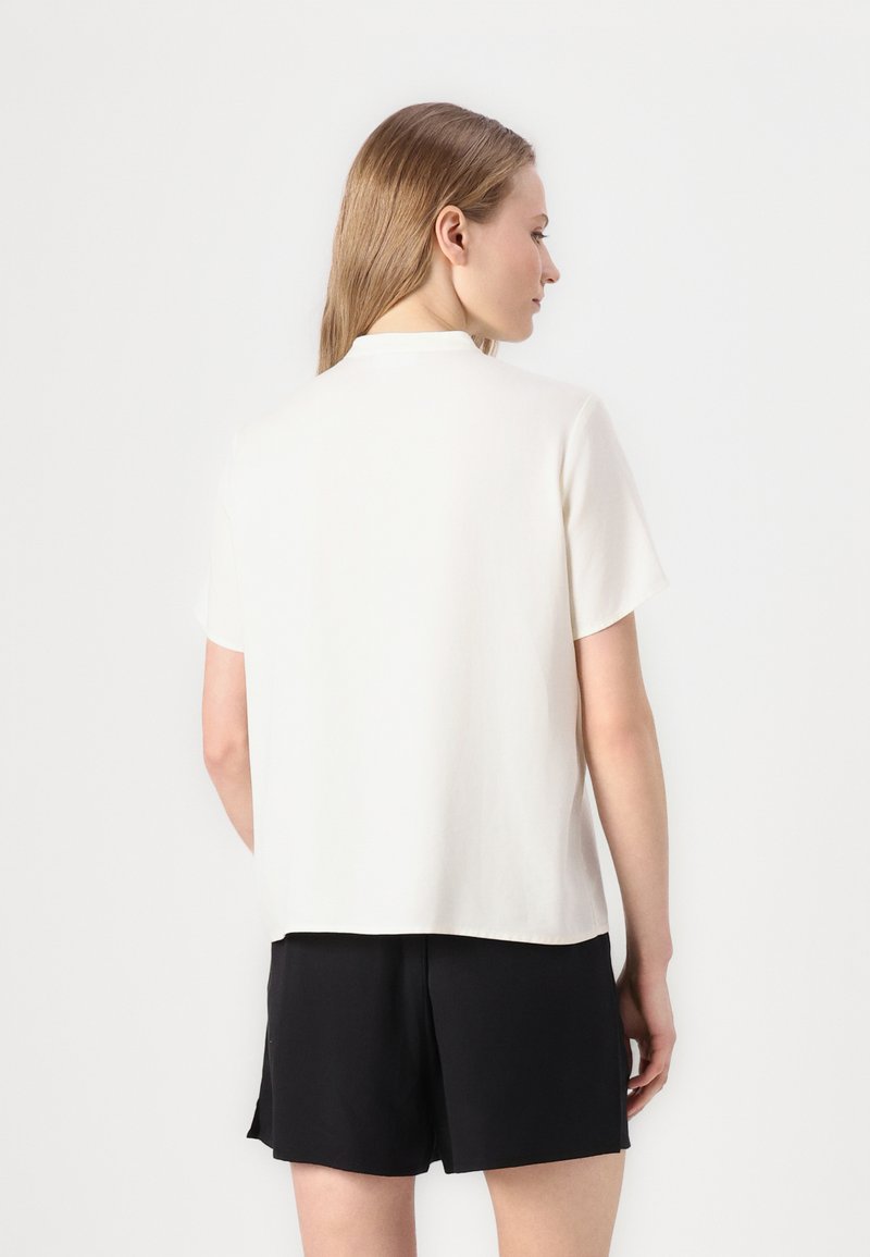 Weißes kurzärmliges Oberteil mit geradem Schnitt und weichem Material, kombiniert mit schwarzen Shorts, von hinten betrachtet.