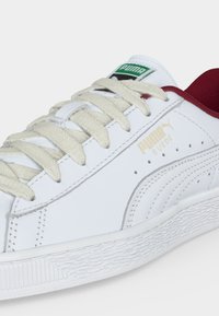 Λευκό σουέντ sneaker Puma με κρεμ κορδόνια, πράσινο λογότυπο στη γλώσσα και κόκκινη εσωτερική επένδυση, εμφανίζεται από κοντινή πλάγια γωνία.