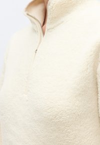 Pulover od flisa krem boje s visokim ovratnikom i poluzatvaračem na patentni zatvarač. Mekana tekstura s udobnim, mekanim izgledom.