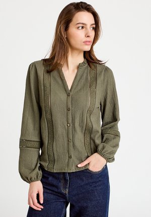 Olivengrøn bluse med tekstureret stof, der har knaplukning foran, stående krave og dekorative udskæringsdetaljer langs sømmene.