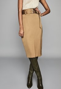 Vrouw in een beige knielange rok met een brede bruine riem, crèmekleurig topje en donkergroene leren laarzen tot aan de knie, staande met één hand op de heup.