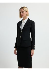 Blazer negro con botones dorados, corte ajustado, dos bolsillos delanteros, combinado con una falda lápiz negra, estilizado sobre una camisa blanca con cuello.