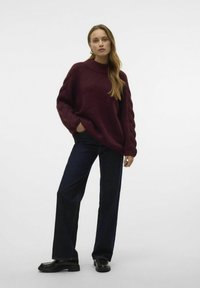 Burgunderfarbener Strickpullover mit hohem Kragen und Kabelstrickärmeln, kombiniert mit dunkelblauen weit geschnittenen Jeans und schwarzen Loafers.