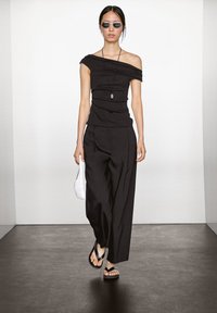 Zwarte off-shoulder top met geplooide textuur, brede zwarte broek en zwarte flip-flops. Houdt een witte gevlochten clutch vast. Minimalistisch ontwerp.