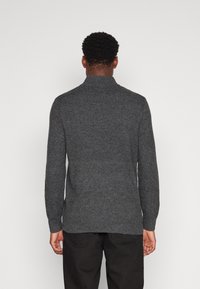 Mann in einem dunkelgrauen Strickpullover und schwarzen Hosen, der mit dem Rücken zur Kamera vor einem schlichten hellen Hintergrund steht.