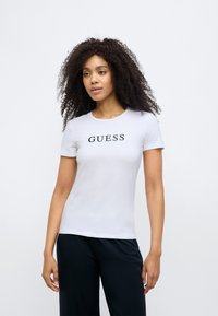 White cotton t-shirt with short sleeves featuring the black logo "GUESS" centered on the chest. Smooth texture, fitted design.