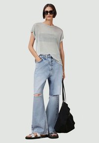 Top cinza claro de mangas curtas, ajuste solto; jeans largos e largados em azul claro; sandálias pretas; mala tote preta; óculos de sol.
