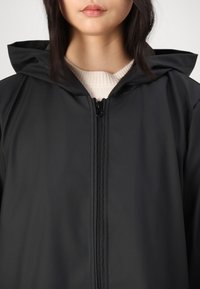 Giacca nera impermeabile con cappuccio, dotata di zip frontale a tutta lunghezza e una texture liscia e opaca. Indossata sopra un maglione leggero.