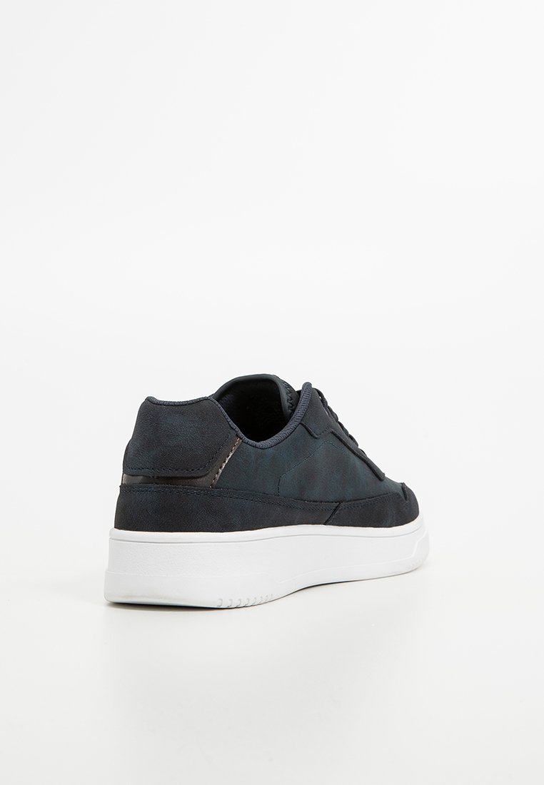 Zapatilla de ante azul oscuro con una textura suave, suela de goma blanca, forma redondeada, parte delantera con cordones y un sutil detalle de costura.