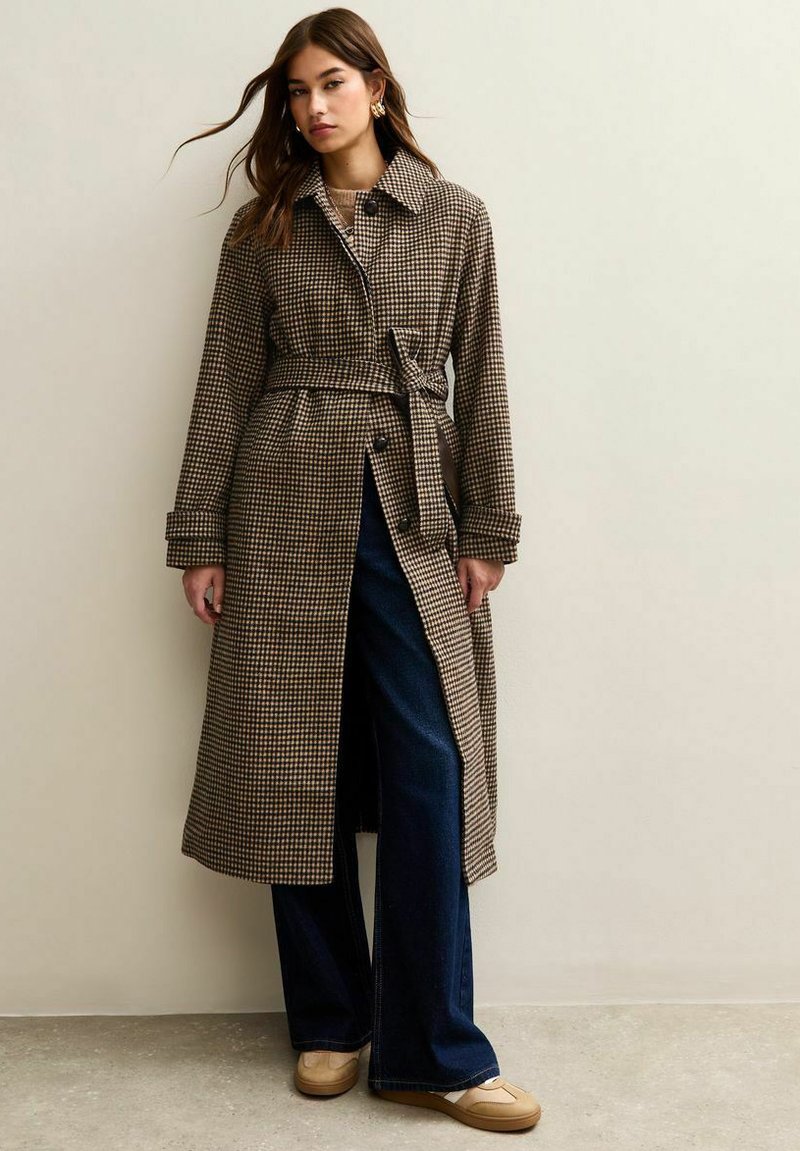 Abrigo trench de cuadros marrones y crema con cinturón en la cintura, mangas largas y cierre de botones, combinado con pantalones de mezclilla oscuros de pierna ancha y zapatillas.