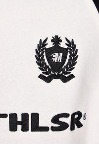 Fekete hímzett logó fehér szöveten, mely pajzsot ábrázol babérlevelekkel és az "M" betűvel, valamint stilizált "ATHLSR®" feliratot.