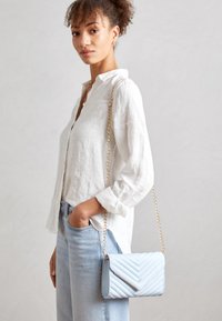 Sac bandoulière matelassé bleu clair avec une sangle chaîne dorée, doté d'un rabat à fermeture et d'un motif chevron. Porté sur une chemise en lin blanche.
