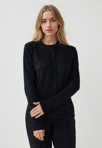 Schwarze Strickjacke aus weichem Stoff, mit strukturierten floralem Applikationen an den Schultern, Knopfverschluss und gerippten Bündchen und Saum.