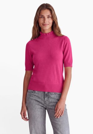 Femme portant un pull rose vif à manches courtes avec un col à volants et un jean gris, debout devant un fond blanc.