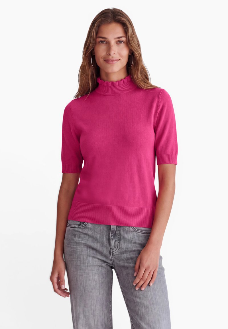 Femme portant un pull rose vif à manches courtes avec un col à volants et un jean gris, debout devant un fond blanc.