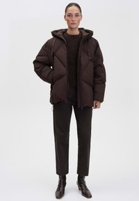 Giacca puffy marrone con cappuccio, caratterizzata da un design trapuntato e chiusura con zip. Indossata sopra un maglione scuro e abbinata a pantaloni scuri e stivali.