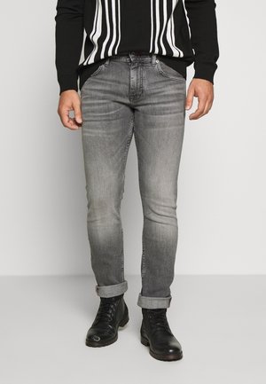 Homme portant un jean gris délavé roulé aux chevilles, des bottes noires à lacets, et un pull noir avec des rayures verticales blanches.