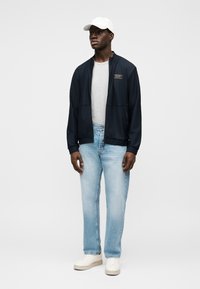 Μπλε σκούρο τζάκετ με φερμουάρ και υφασμάτινο σχέδιο, ανοιχτό γκρι μπλουζάκι, ανοιχτό μπλε τζιν παντελόνι και λευκά αθλητικά παπούτσια, συνοδευόμενα από λευκό καπέλο.