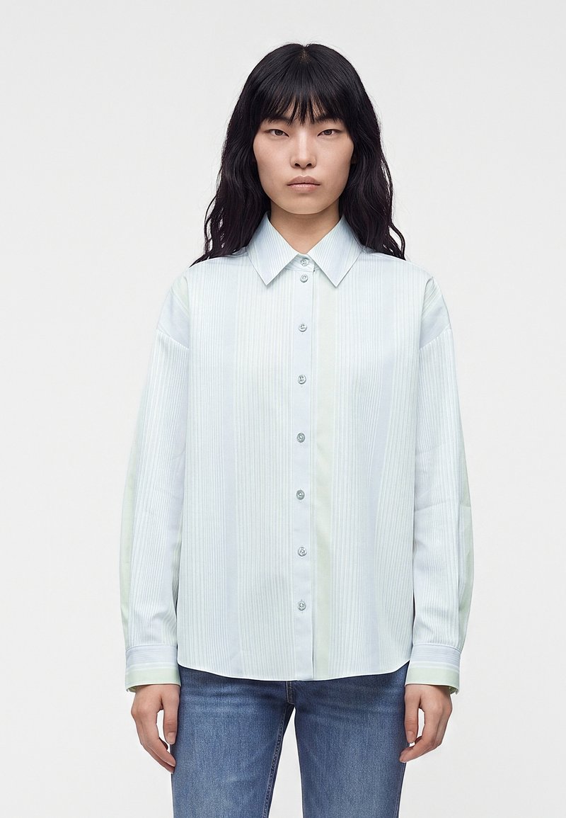 Femme aux longs cheveux noirs porte une chemise à rayures bleu clair avec boutons et un jean bleu, debout sur un fond gris clair uni.