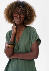 Zielony top z krótkim rękawem i dekoltem w kształcie V. Model nosi duże brązowe okulary przeciwsłoneczne oraz nałożone na siebie bransoletki o różnych fakturach i kolorach.