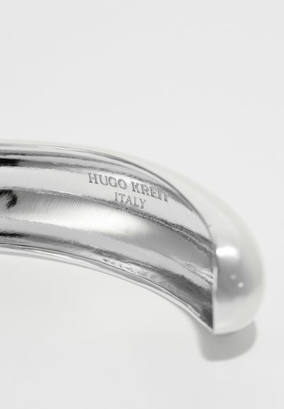 Gepolijste zilveren metalen handgreep met een gebogen ontwerp. Gegraveerde tekst leest "HUGO KREIT ITALIË," en toont een gladde textuur en reflecterend oppervlak.