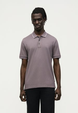 Άνδρας με σκοτεινό δέρμα και dreadlocks, που φοράει κοντομάνικο πουκάμισο πόλο σε μωβ απόχρωση με λογότυπο "BOSS" και μαύρο παντελόνι, στέκεται μπροστά σε μονόχρωμο φόντο.