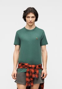 Νεαρό άτομο με κοντά σκούρα μαλλιά φοράει πράσινο κοντομάνικο πουκάμισο με μικρό κεντημένο ελάφι, γκρι σορτς και κόκκινο-μαύρο καρό πουκάμισο δεμένο γύρω από τη μέση.