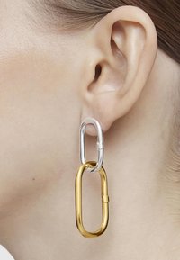 Primer plano de una oreja que lleva un pendiente de dos eslabones con un óvalo superior de plata y un eslabón inferior ovalado más grande de oro.