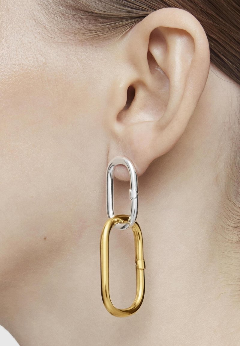 Primer plano de una oreja que lleva un pendiente de dos eslabones con un óvalo superior de plata y un eslabón inferior ovalado más grande de oro.