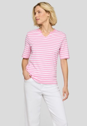 Frau mit kurzen blonden Haaren, die ein rosa-weiß gestreiftes Kurzarmshirt und weiße Hosen trägt, mit einer Hand in der Tasche.