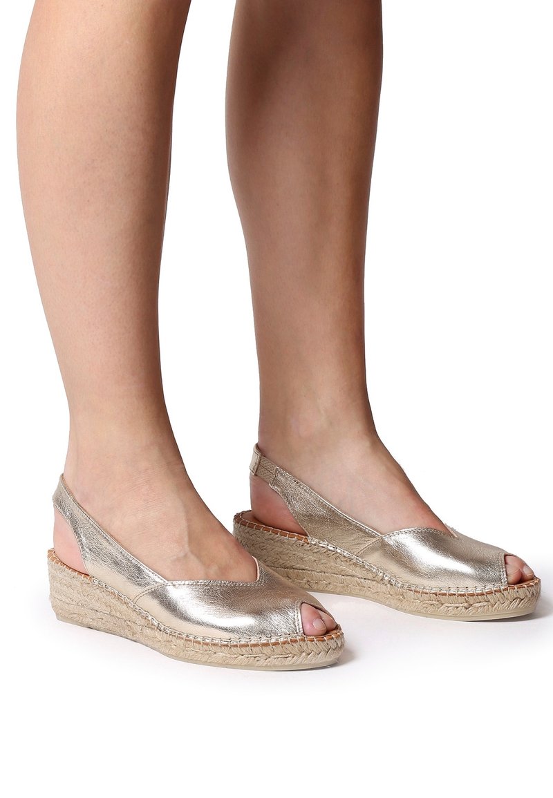 Pieds portant des sandales compensées à brides arrière métalliques dorées, avec orteils découverts et semelles plateformes en jute tressé, sur fond blanc.
