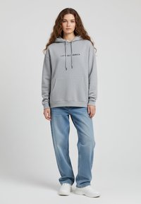 Felpa grigia in cotone con tasca frontale, cappuccio con cordini e testo nero che dice "CONIUGHIAMO!" Abbinata a jeans baggy azzurri chiari e scarpe da ginnastica bianche.