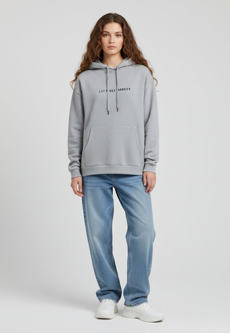 Felpa grigia in cotone con tasca frontale, cappuccio con cordini e testo nero che dice "CONIUGHIAMO!" Abbinata a jeans baggy azzurri chiari e scarpe da ginnastica bianche.
