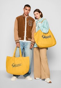Kollase kangast kottidega, millel on "Gant" logo; üks inimene kannab pruuni kordjakki, teine aga triibulist särki koos beežide pükstega.