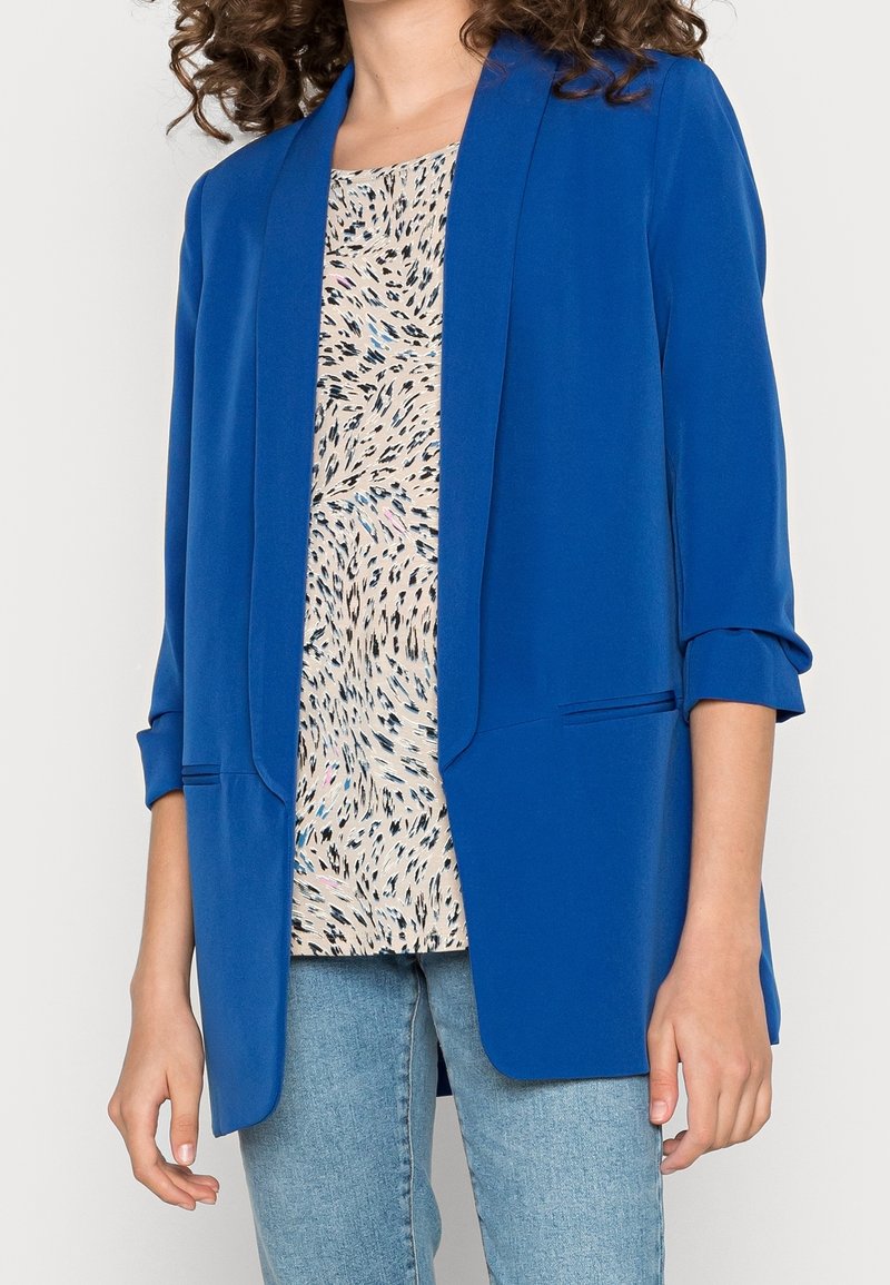 Vrouw met een felblauwe blazer met opgerolde mouwen over een beige blouse met patroon en lichtblauwe spijkerbroek, staand tegen een effen achtergrond.