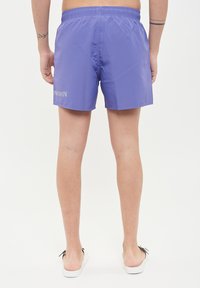 Pantaloni da bagno viola con vita elasticizzata, texture liscia e logo bianco stampato sul retro. Lunghezza corta e design minimal.