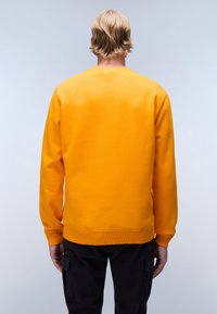 Felpa gialla con polsini e orlo a costine. Materiale misto cotone, design a collo rotondo, texture liscia, indossata con pantaloni cargo scuri.