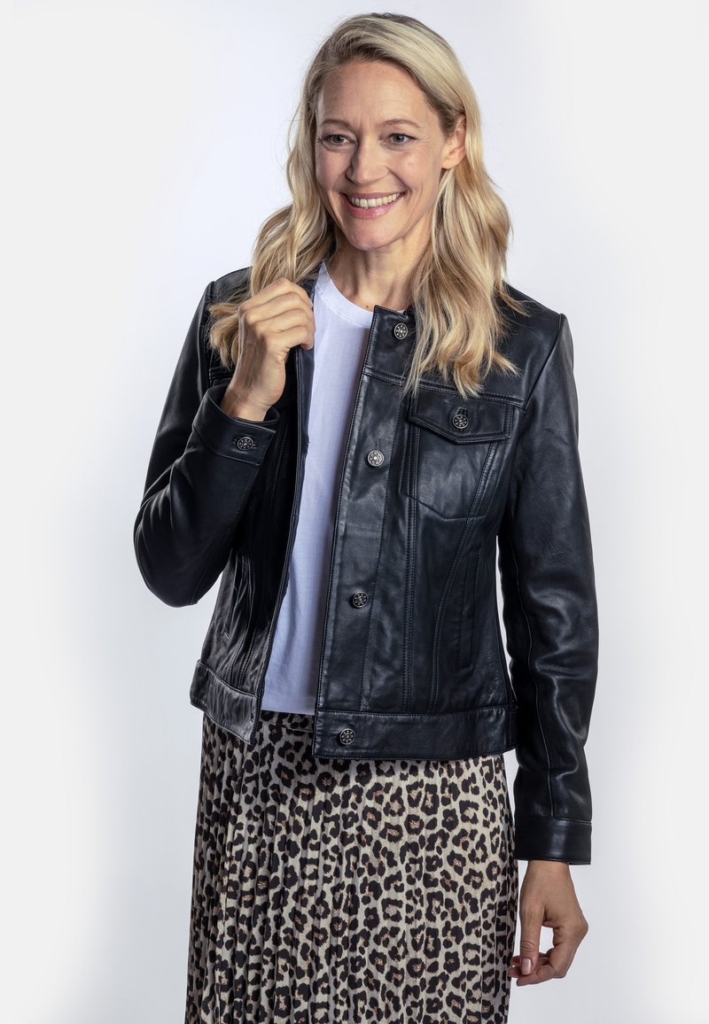 Schwarze Lederjacke mit Knopfverschluss, seitlichen Taschen und strukturiertem Design, getragen über einem weißen Hemd und einem Leopardenmuster-Rock.