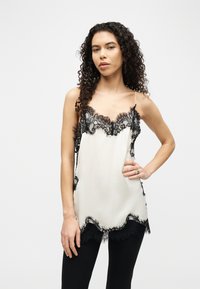 Γυναίκα με σγουρά μαλλιά, φοράει λευκό αμάνικο μεταξωτό τοπ με μαύρη δαντέλα στα τελειώματα και μαύρο παντελόνι, ποζάροντας με το χέρι στη μέση μπροστά σε λευκό φόντο.