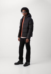 Czarna, pikowana kurtka z kapturem, pomarańczowa koszula noszona na wierzchu, czarne spodnie i czarne obuwie, na jednolitym tle.