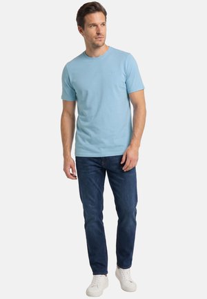 Mand stående iført lyseblå kortærmet t-shirt, mørkeblå jeans og hvide sneakers, kigger let til siden.