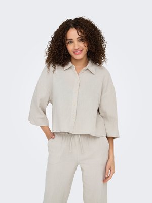 Donna con capelli ricci che sorride, indossa una camicia beige larga con bottoni e maniche a tre quarti e pantaloni coordinati con coulisse, mani nelle tasche.