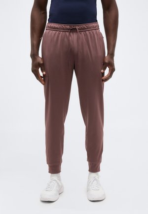 Mand iført mauve joggingbukser, hvide sneakers og sokker, stående mod en ensfarvet lys baggrund med hænderne afslappet ned langs siderne.