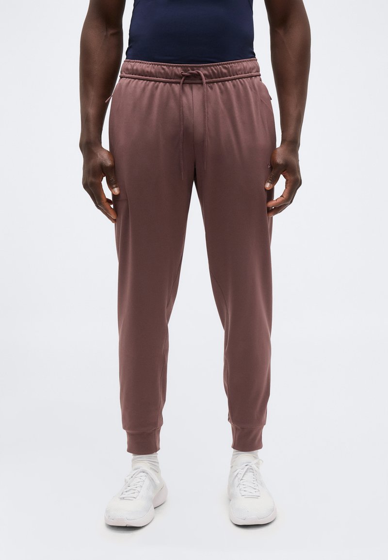 Άνδρας που φοράει μωβ αθλητική φόρμα, λευκά αθλητικά παπούτσια και κάλτσες, στέκεται απέναντι σε ένα απλό φωτεινό φόντο με τα χέρια χαλαρά δίπλα στο σώμα.